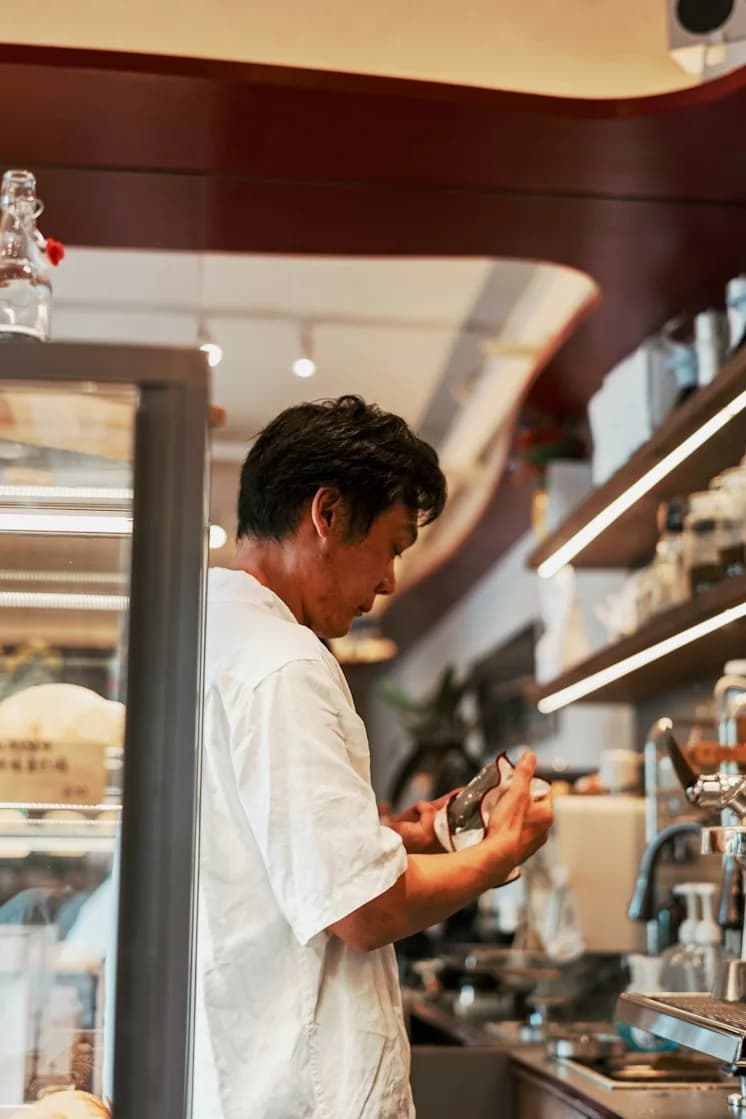 A cook in a white shirt at the pass of a warmly lit restaurant, looking down at his hands
