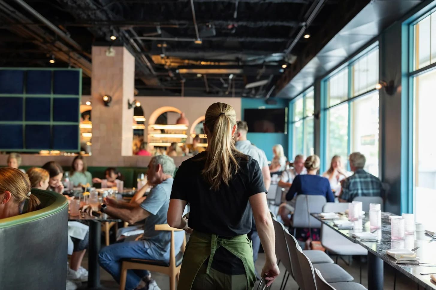 A server with a ponytail walking between busy tables in a bright restaurant with blue walls