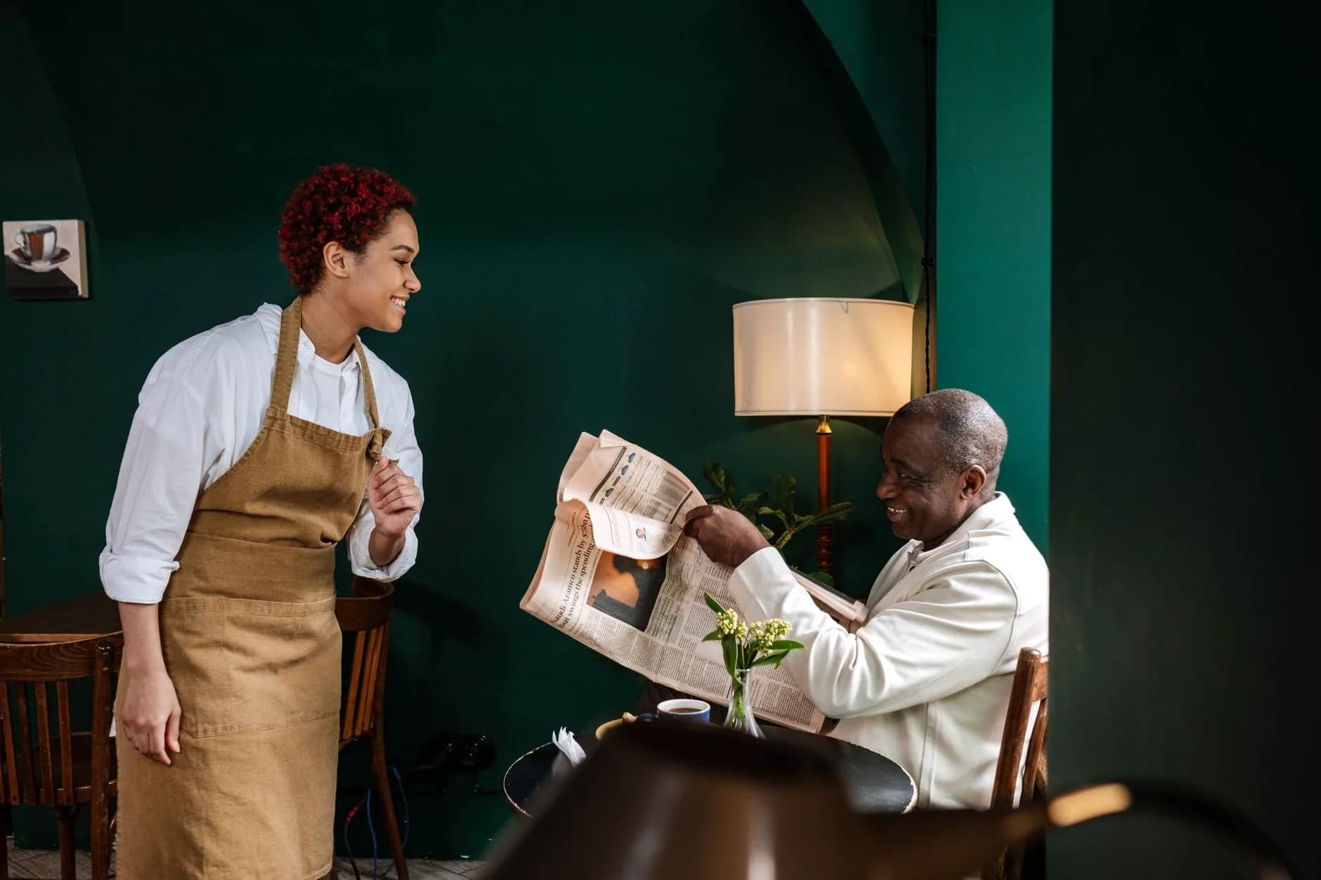 A server in a linen apron laughing with a guest who reads a newspaper at his table, in a dark green dining room