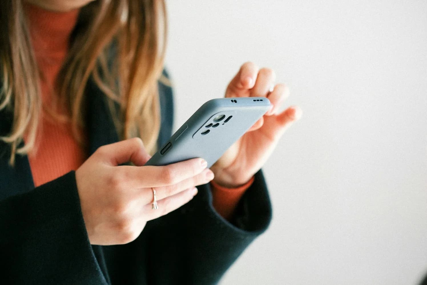 A woman in a dark coat and an orange jumper holding a pale blue phone in both hands