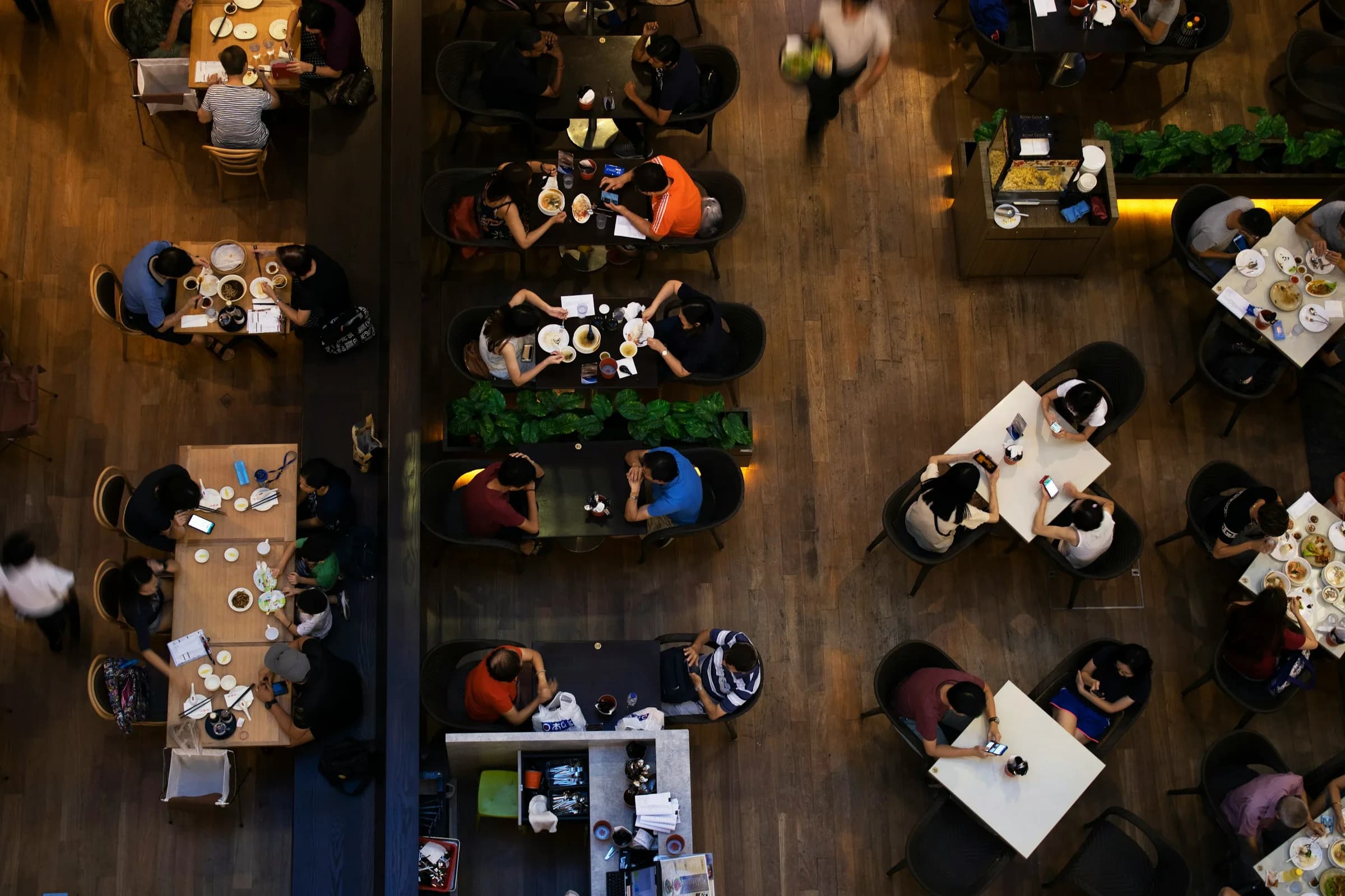 A restaurant floor seen from above: guests at wooden tables and a waiter walking between them