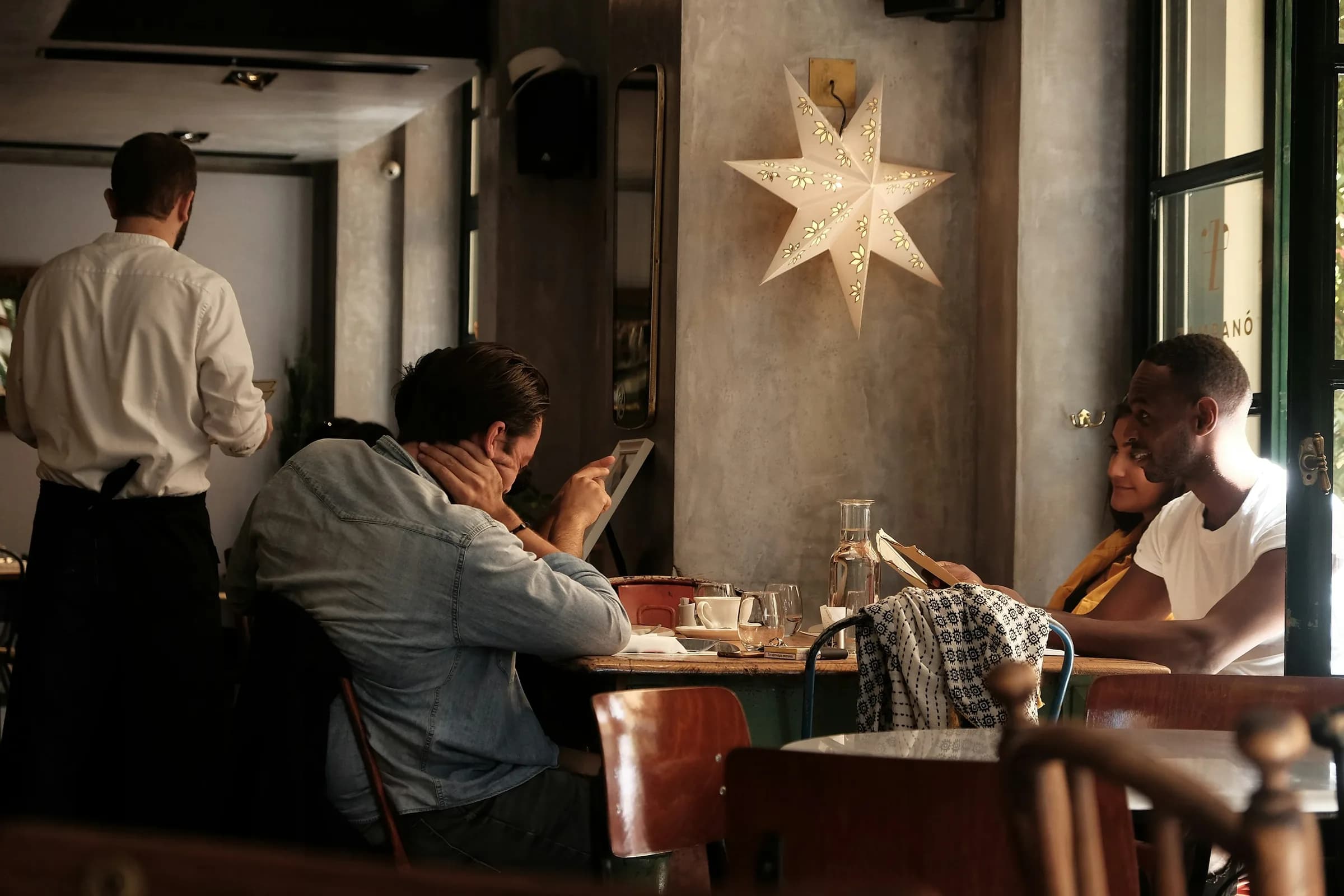 A café with a paper star lamp: a waiter in a white shirt and guests reading at their tables in warm light