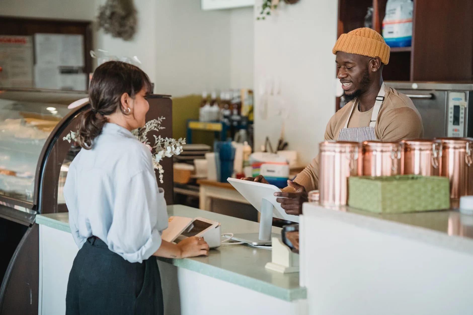 A guest in a light blue shirt at a café counter, the barista smiling behind the pastry case