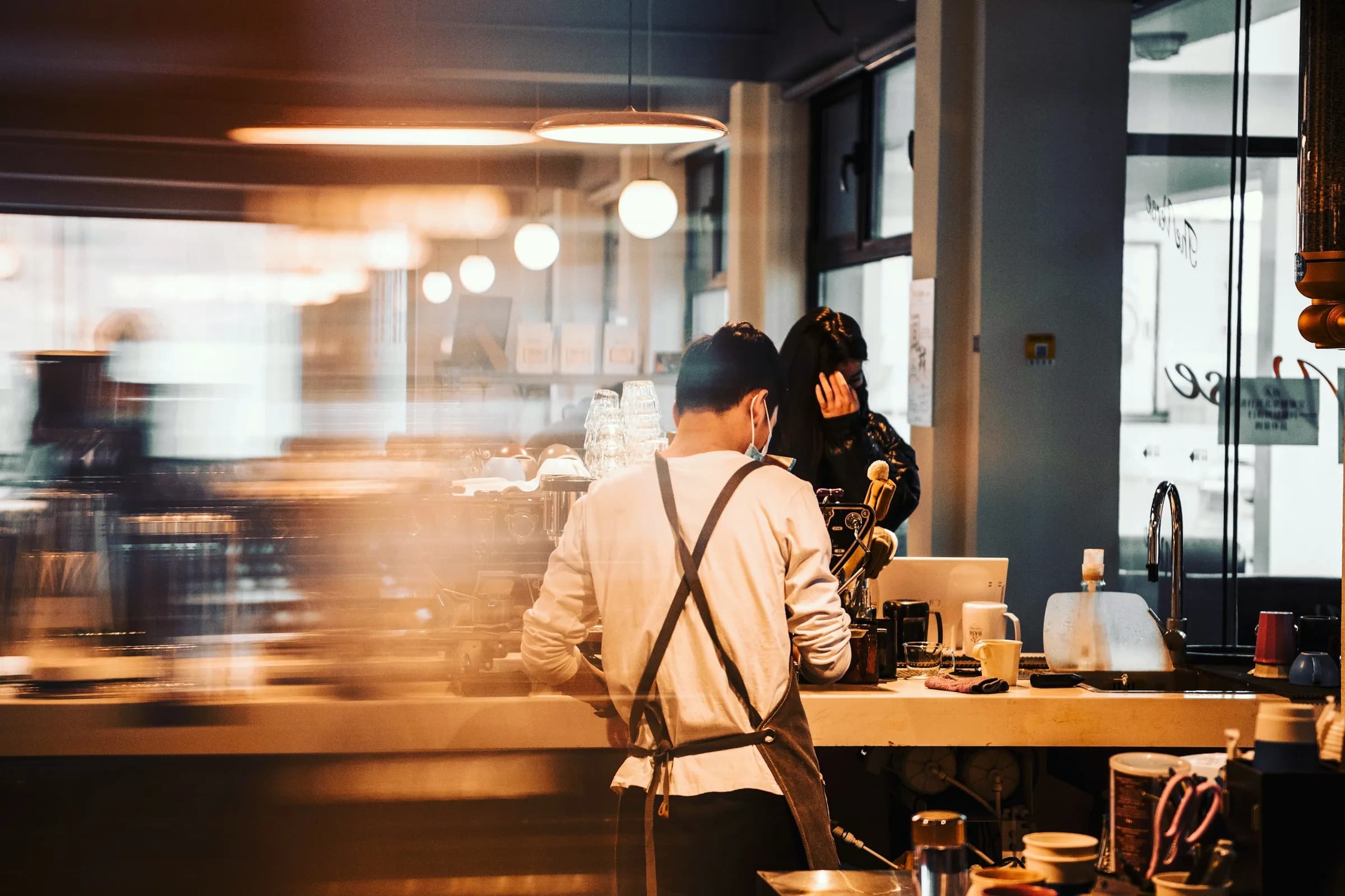 A barista in an apron working at a café counter, the room behind blurred with movement