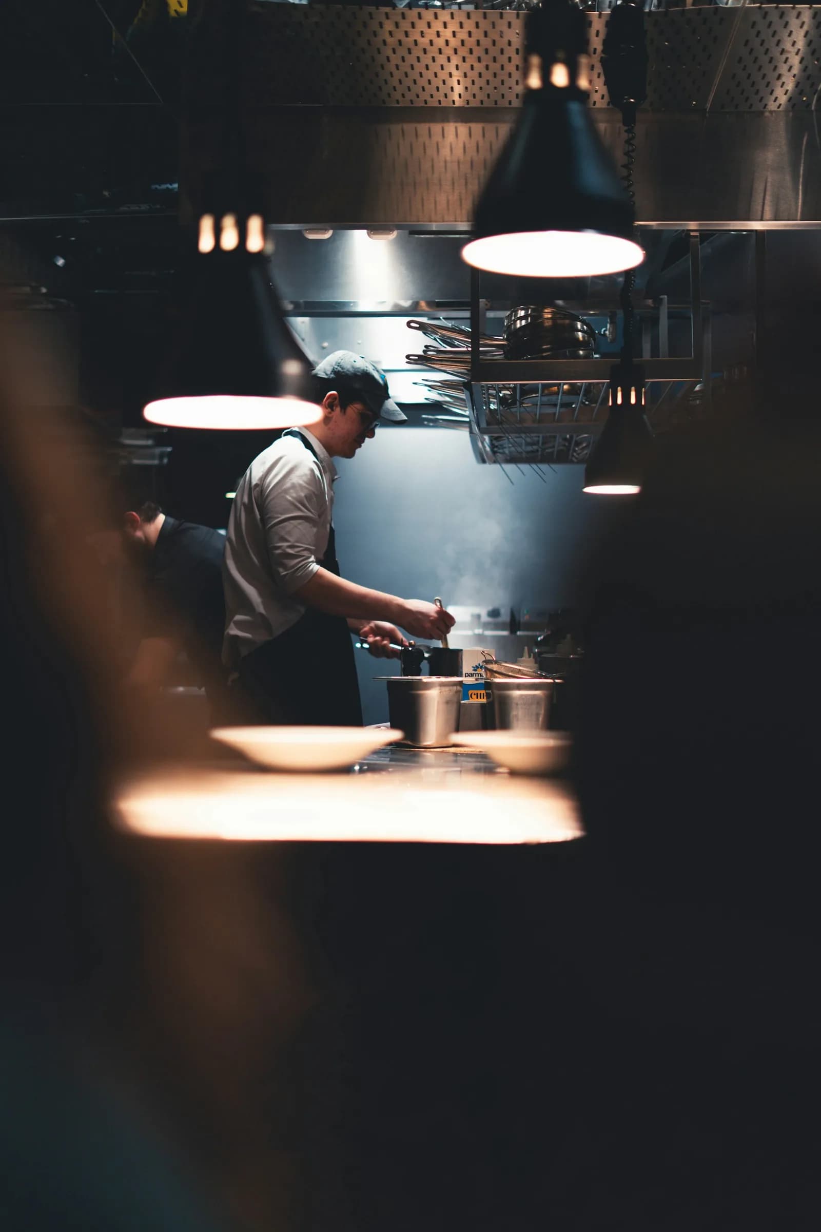A cook in a cap stirring a pot at the stove of a dark restaurant kitchen, under a warm pendant lamp