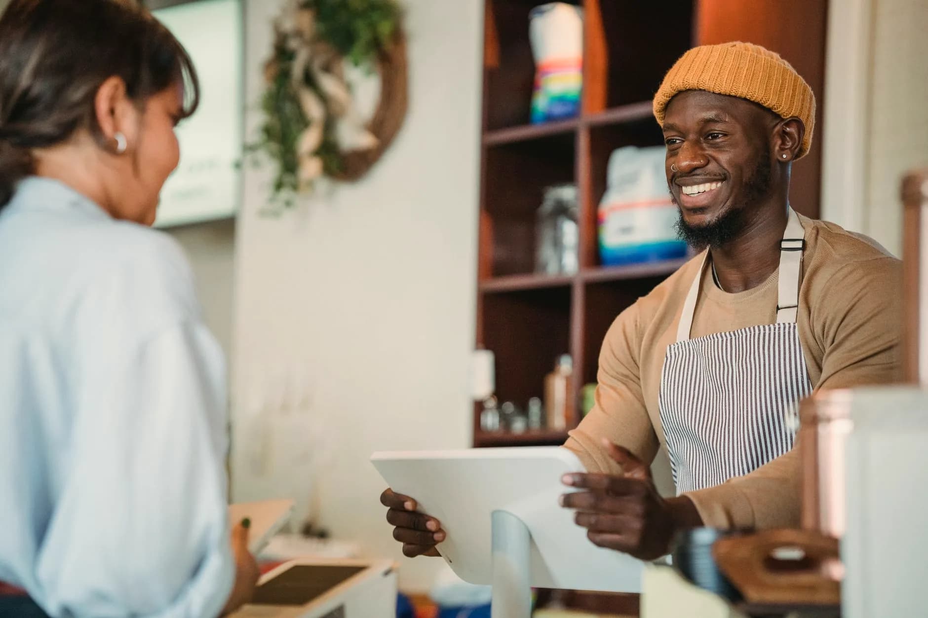 A smiling counter worker in a beanie and striped apron hands a tablet across the counter to a guest