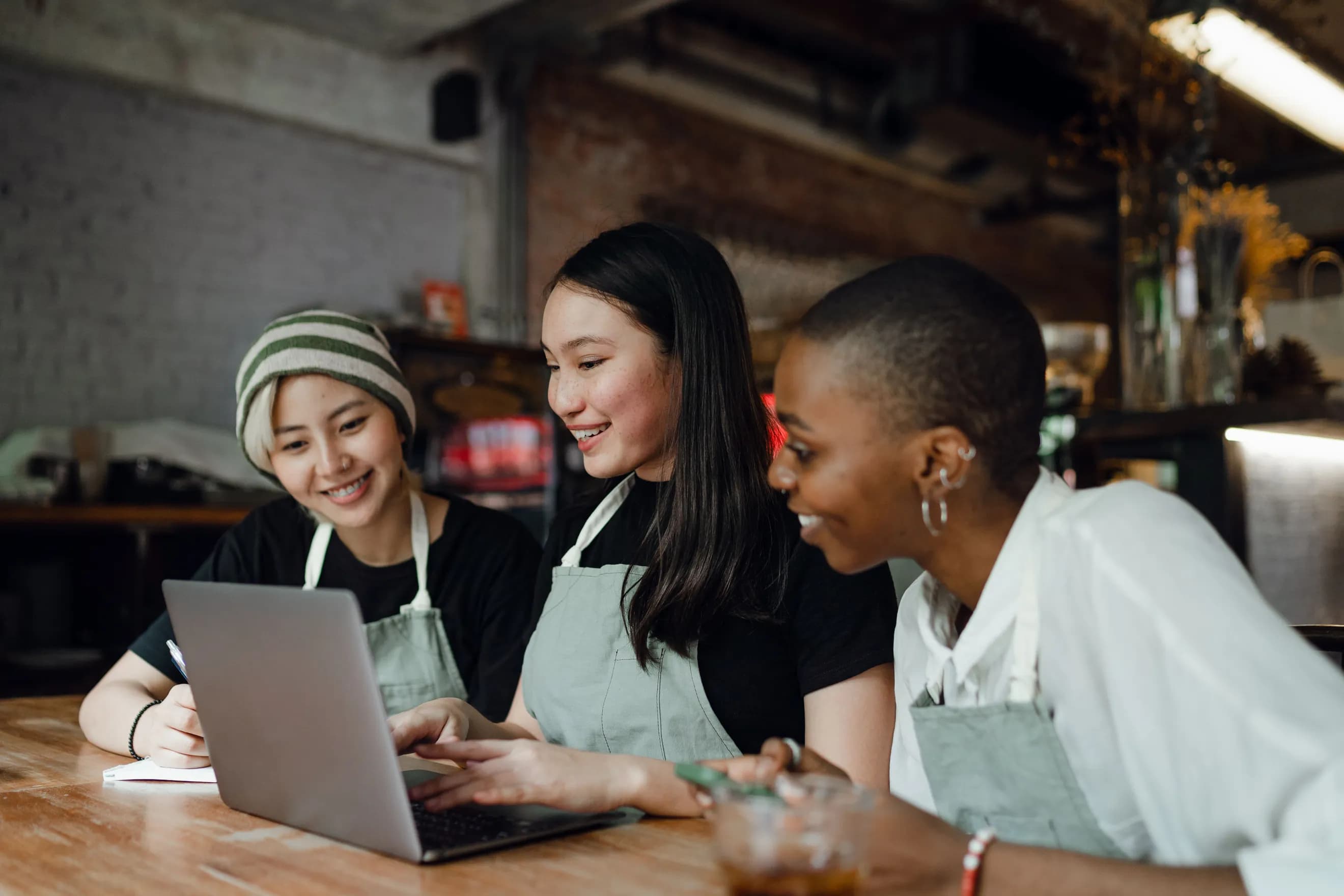 Three restaurant team members in aprons laughing together around a laptop at a wooden table