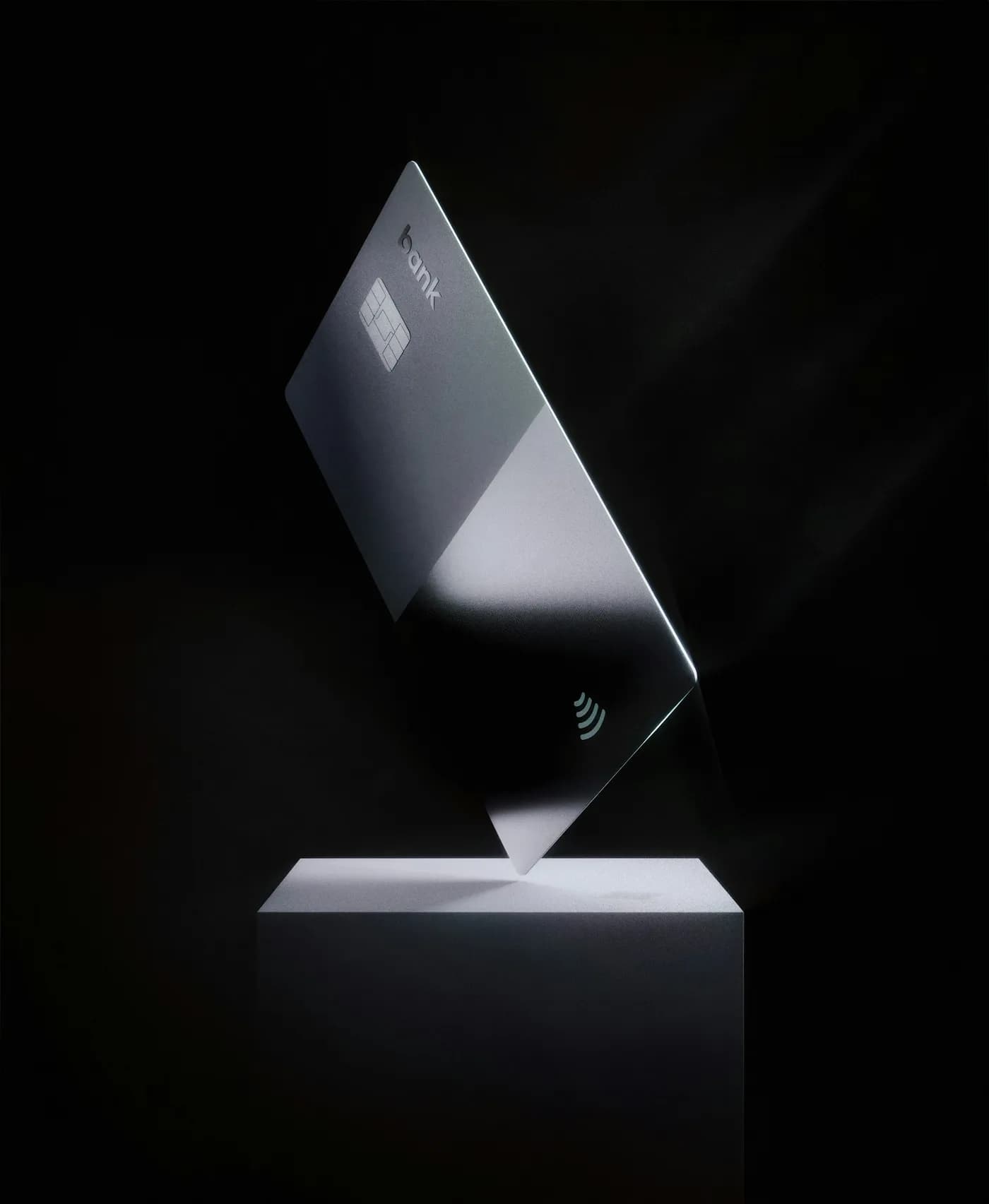 A silver contactless bank card balanced on its corner on a white plinth, lit against a black background