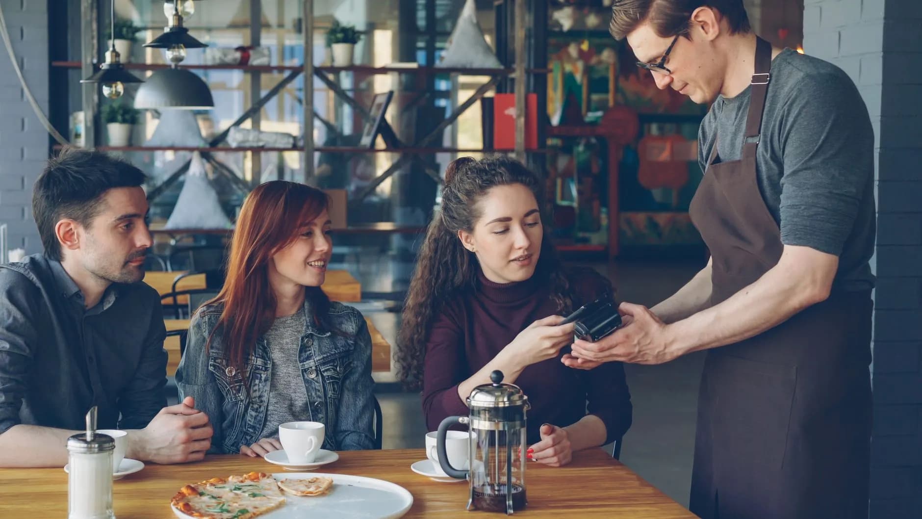 A waiter in a brown apron taking a guest’s payment at a café table while her friends look on over coffee